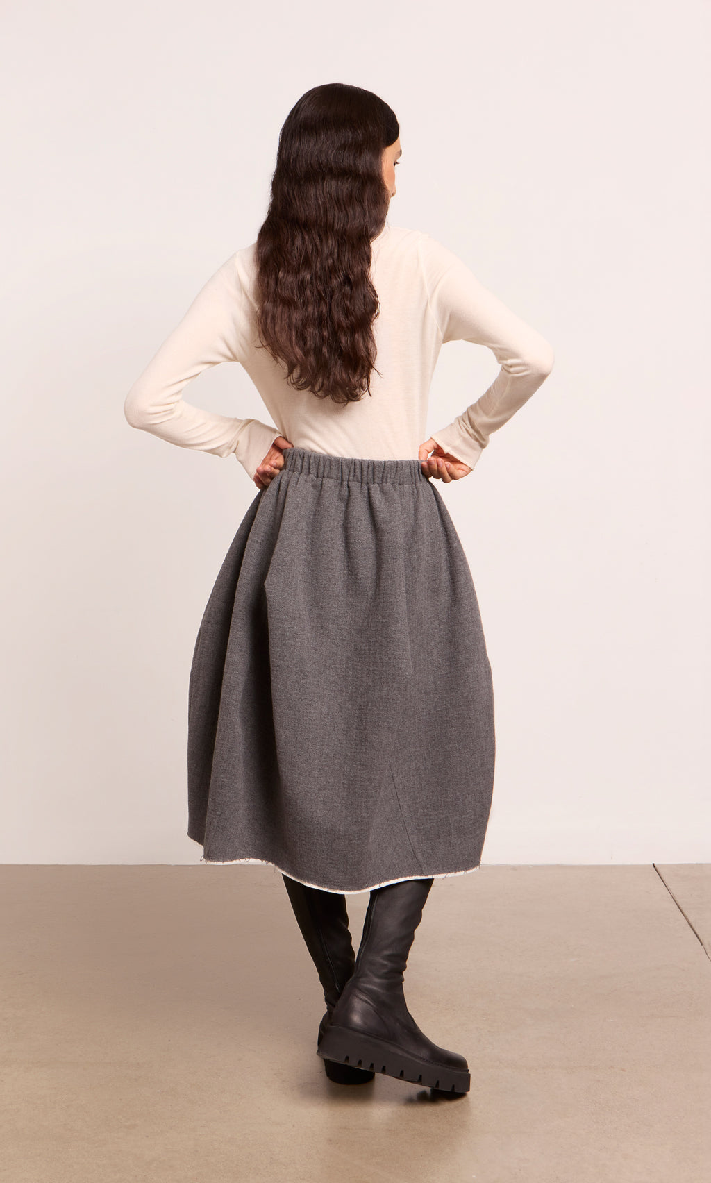 Plümo  Woman stands hands on hips in a Ghent Skirt, cream top, and black boots, facing away from the camera.