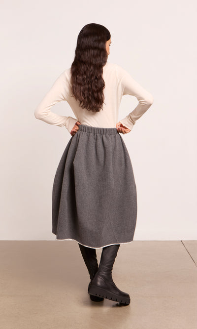 Plümo  Woman stands hands on hips in a Ghent Skirt, cream top, and black boots, facing away from the camera.