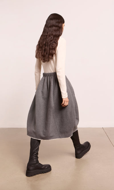 Plümo  Person in a white top, gray skirt, and Long Prato Boots walks on a beige floor, facing away.