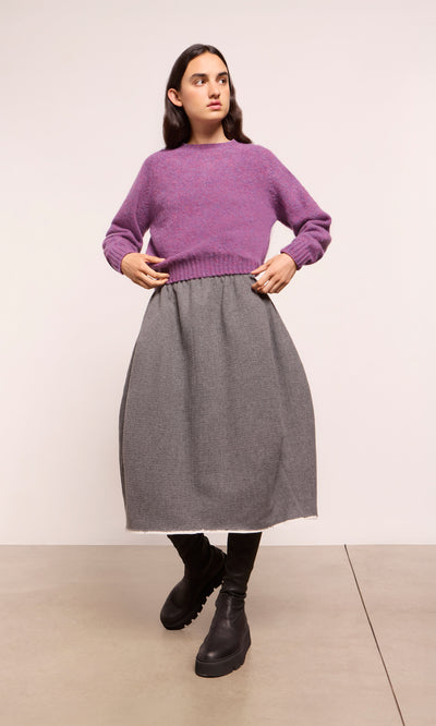 Plümo  Woman in a purple sweater and Ghent Skirt stands on concrete floor, wearing black knee-high boots.