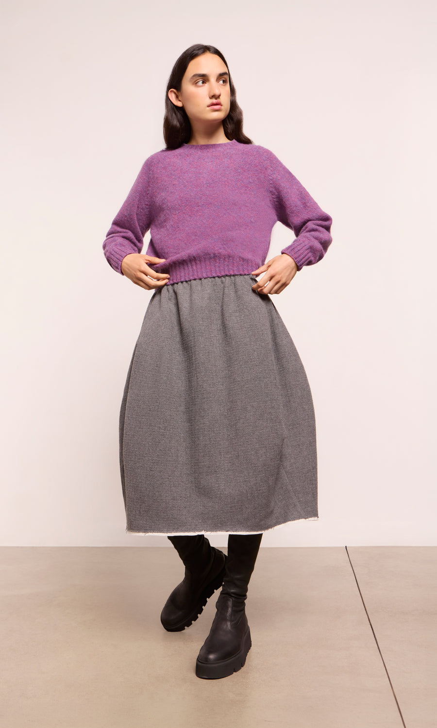 Plümo  Woman in a purple sweater and Ghent Skirt stands on concrete floor, wearing black knee-high boots.