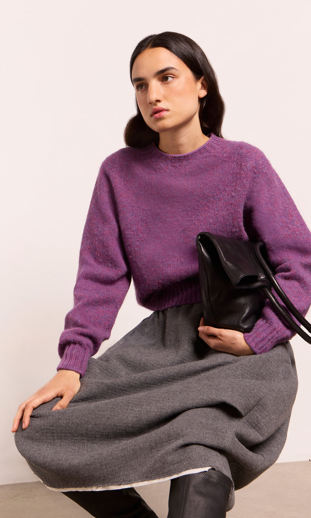 Plümo  Woman in a purple sweater and Ghent Skirt sits with a black bag, looking away with a neutral expression.