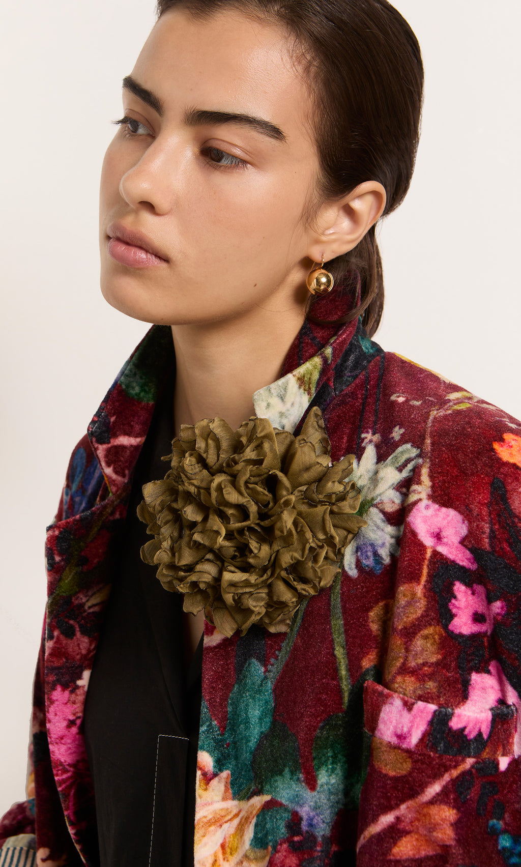 Plümo  Woman in a Paris atelier wears the Velvetine Blazer, adorned with a bold green flower pin and gold earring, gazing aside.