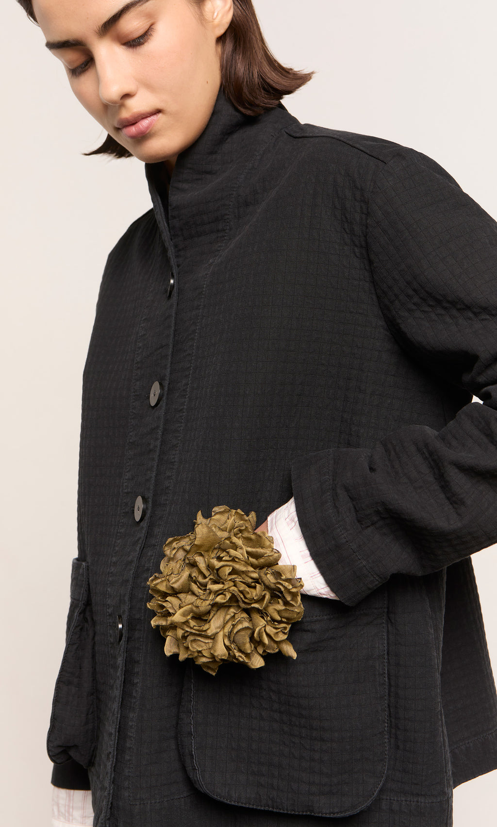Plümo  Person in a black jacket with a Peony Corsage, an oversized olive pleated floral brooch, on one pocket.