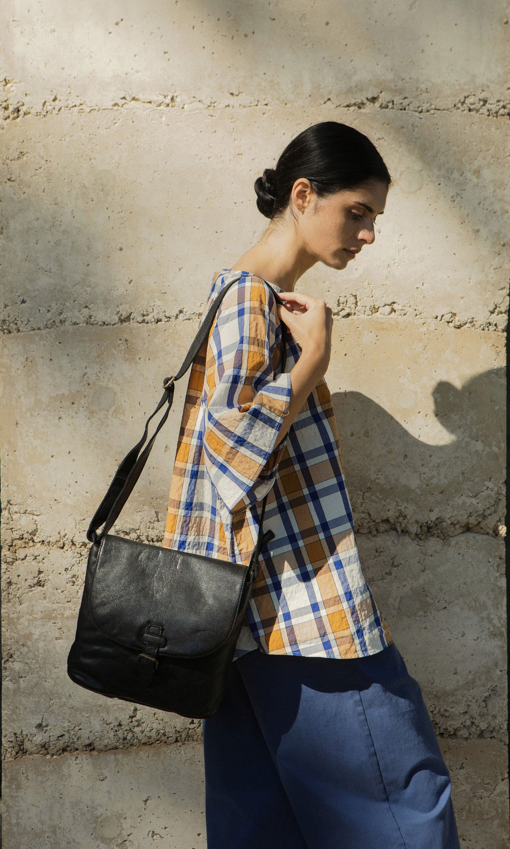 Plümo- Woman in plaid shirt and blue pants stands sideways with the Beck Crossbody Bag against a beige wall.