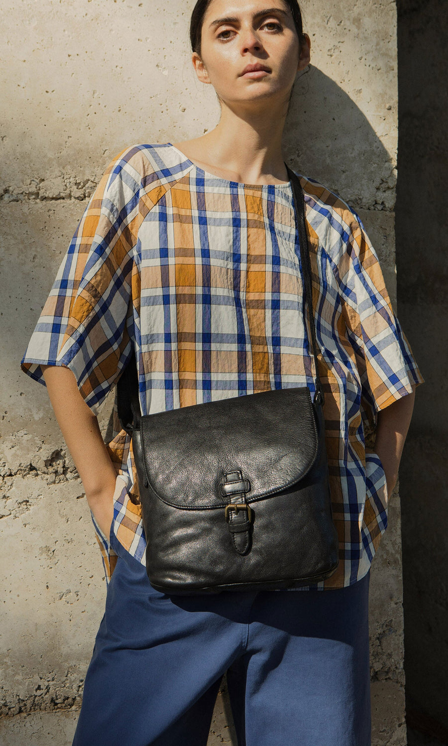 Plümo- Woman in a plaid shirt and blue pants carries a Beck Crossbody Bag with adjustable strap by a stone wall.