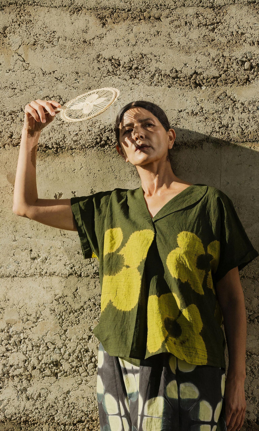 Plümo- Person standing against a textured wall, hiding their face with the Hanoi Fan - Natural, wearing a green floral shirt.