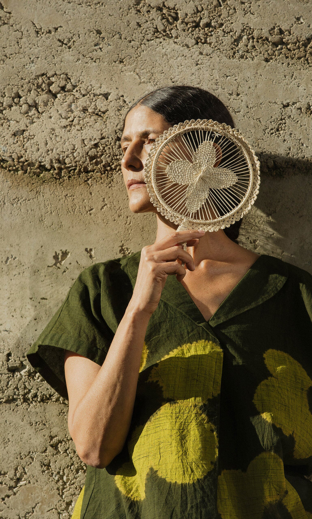 Plümo- A woman in a green dress holds a Hanoi Fan - Natural, partially covering her face against a textured wall.
