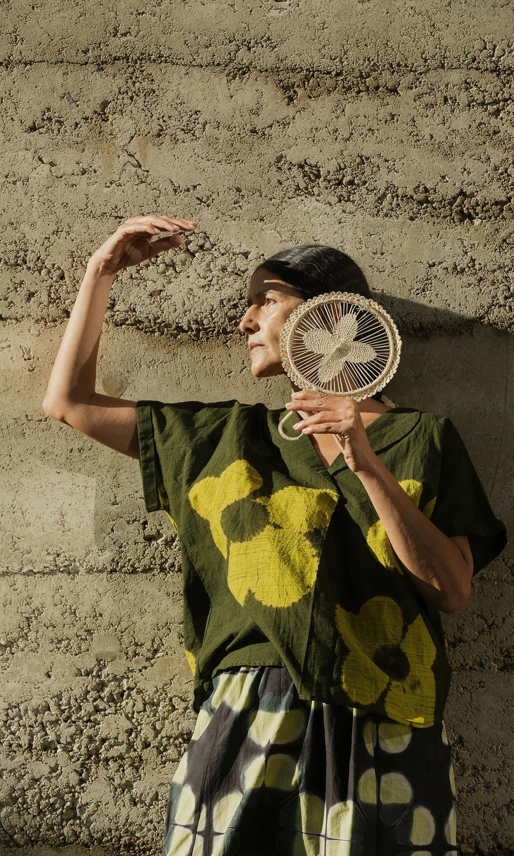 Plümo- Person in a green floral shirt poses with the Hanoi Fan - Natural, covering their face and raising one arm by a textured wall.