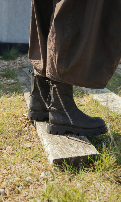 Plümo- Wearing artisan-crafted Harrison Boots, standing on a wooden plank outdoors amid grass and rocks.