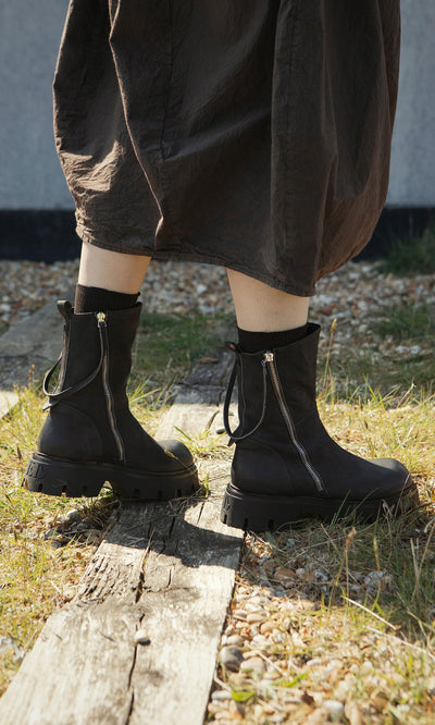 Plümo- Person in a brown skirt and Harrison Boots stands on grass and wooden planks outdoors.
