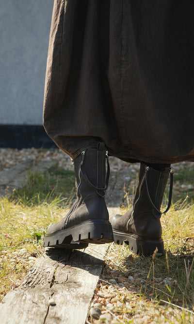 Plümo- Person stands outdoors in tall grass on a wooden plank, wearing loose dark pants and artisan-crafted Harrison Boots.