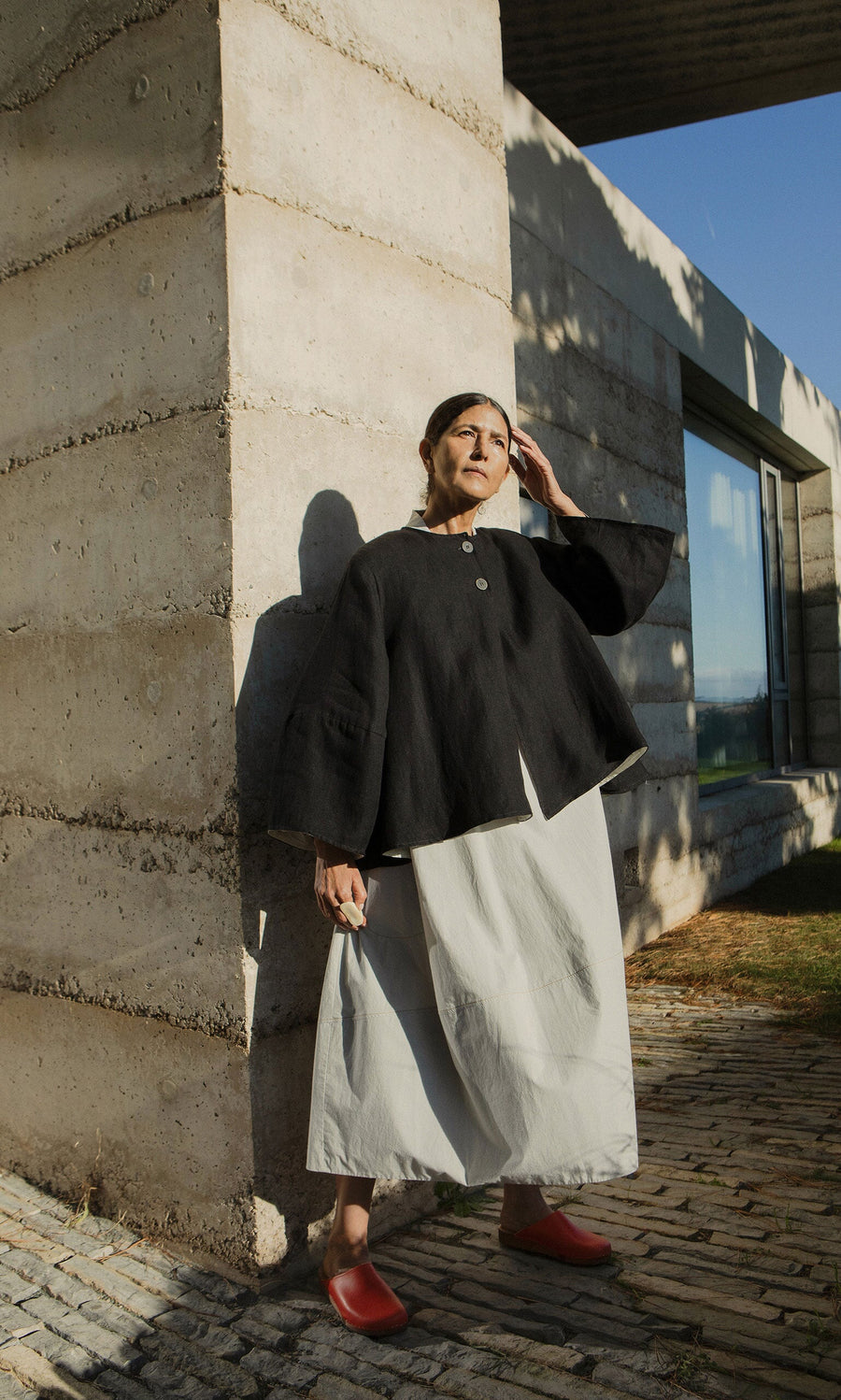 Plümo- Woman in Holden Jacket standing by concrete building, in sunlight, wearing red shoes and a white skirt.
