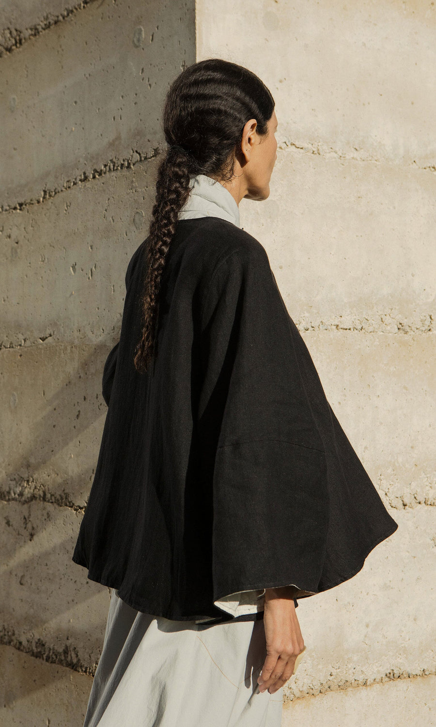 Plümo- Person with long braided hair in a loose Holden Jacket stands sideways by a textured concrete wall.
