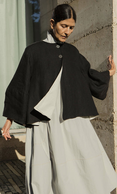 Plümo- A person wearing the black Holden Jacket over a light dress stands by a concrete wall in natural sunlight.