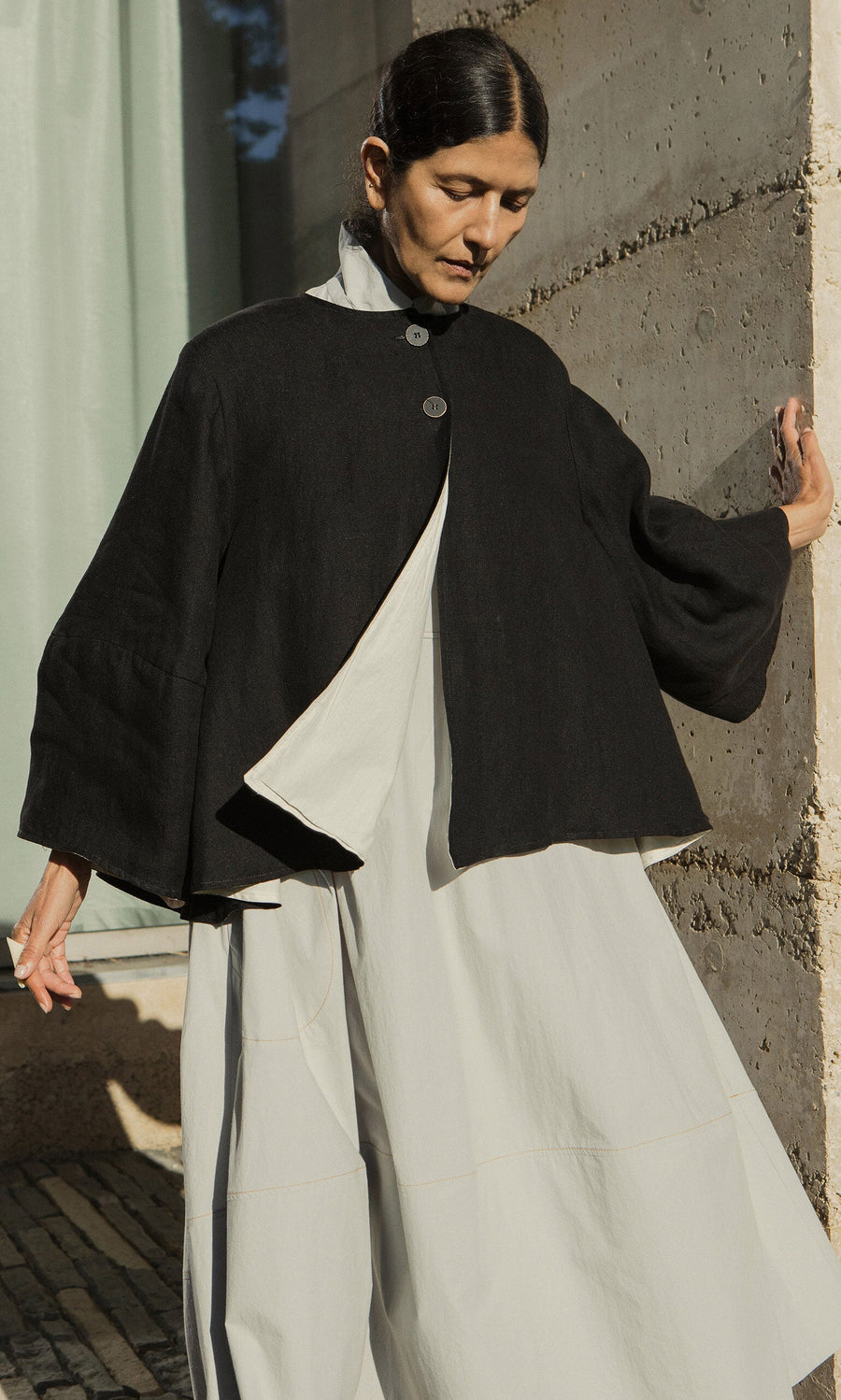 Plümo- A person wearing the black Holden Jacket over a light dress stands by a concrete wall in natural sunlight.