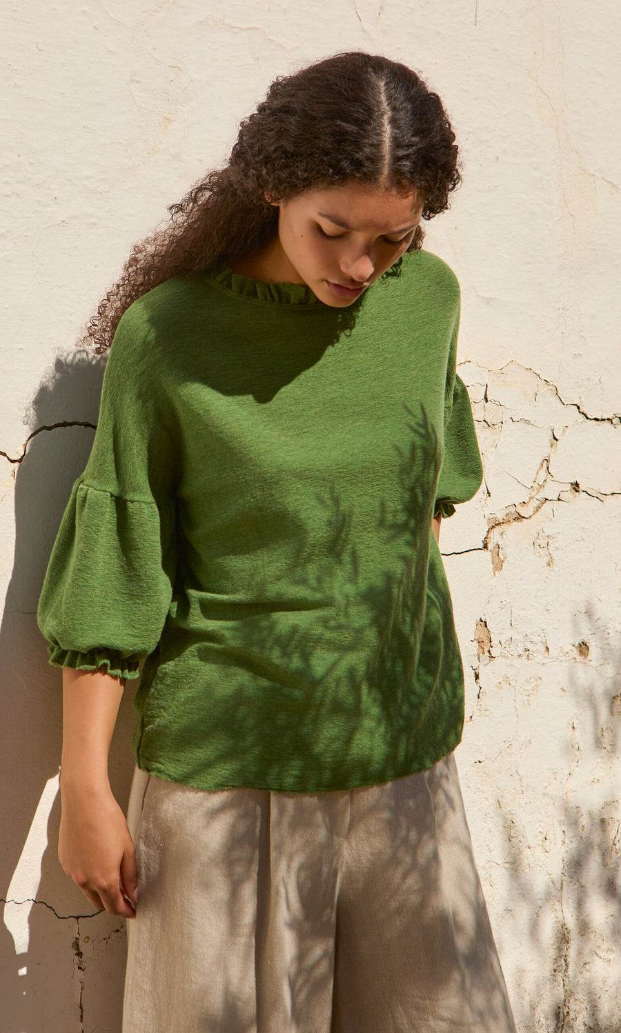 Plümo- Woman in the Hopper Blouse stands by a cracked wall, leafy shadows falling across her Danish design top and beige pants.