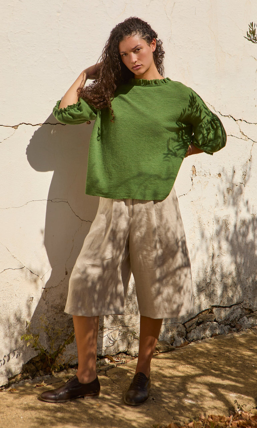 Plümo- Woman in a Hopper Blouse and beige culottes stands by a sunlit white wall with leaf shadows on her.
