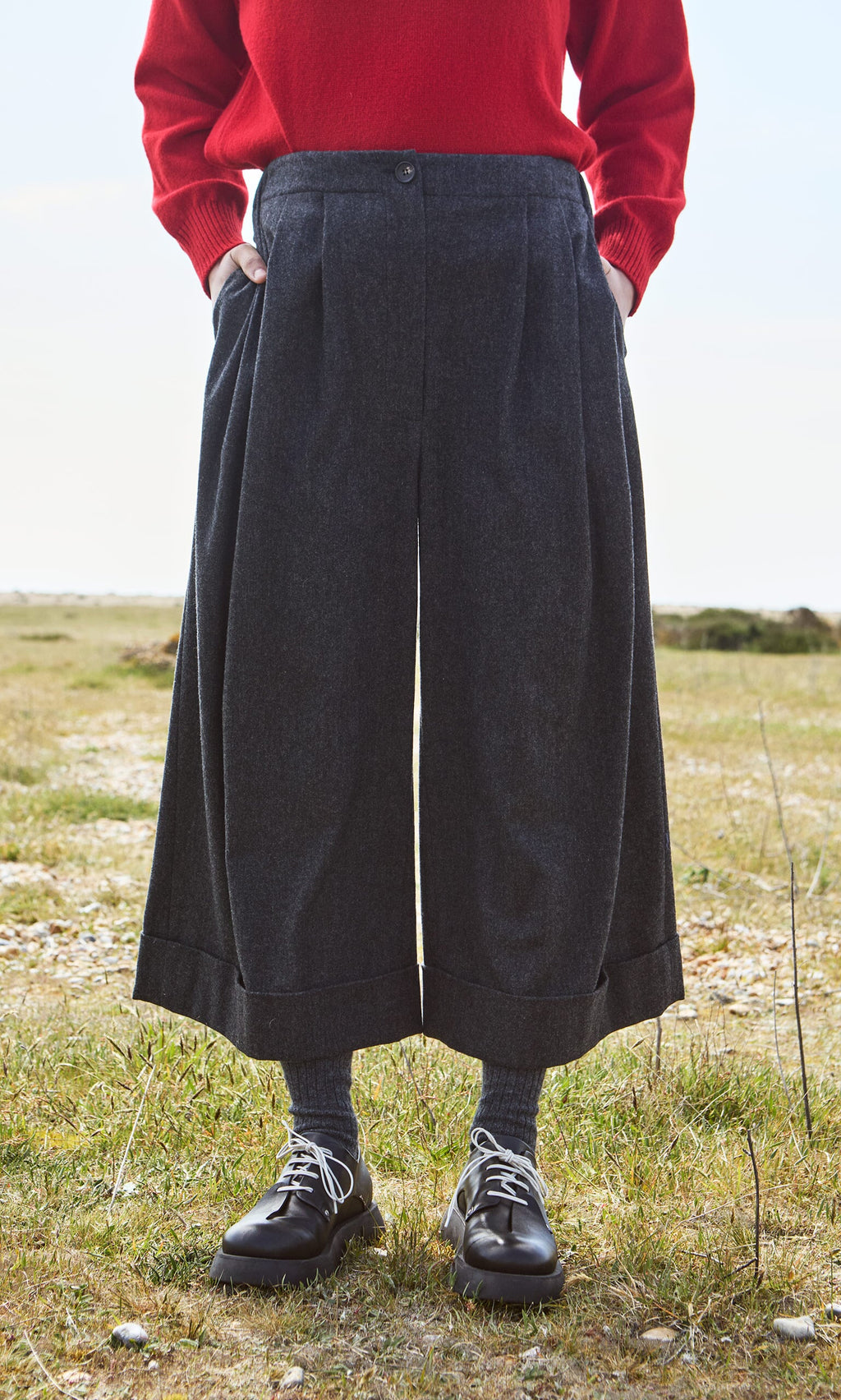 Plümo- Person wearing Horton Pants, a red sweater, gray socks, and black shoes stands outdoors on the grass.