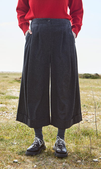 Plümo- Person wearing Horton Pants, a red sweater, gray socks, and black shoes stands outdoors on the grass.
