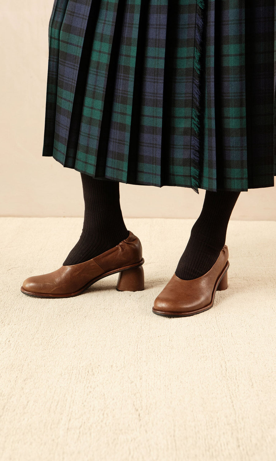 Plümo- Person wearing Solene Heels, a green and blue plaid skirt, and black socks stands on a cream carpet.
