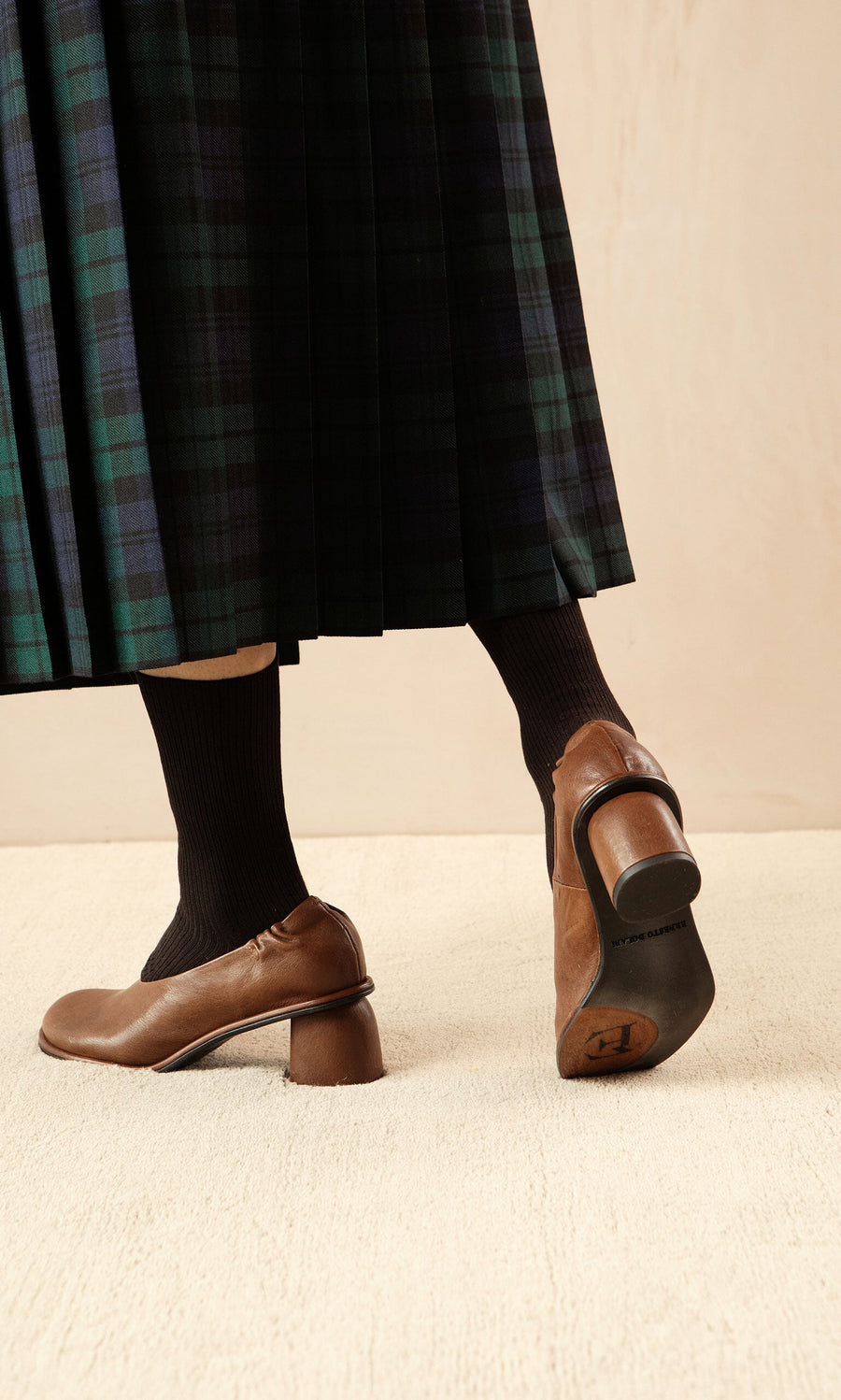 Plümo- Person wearing a green plaid skirt, black socks, and Solene Heels walks on a light carpeted floor.