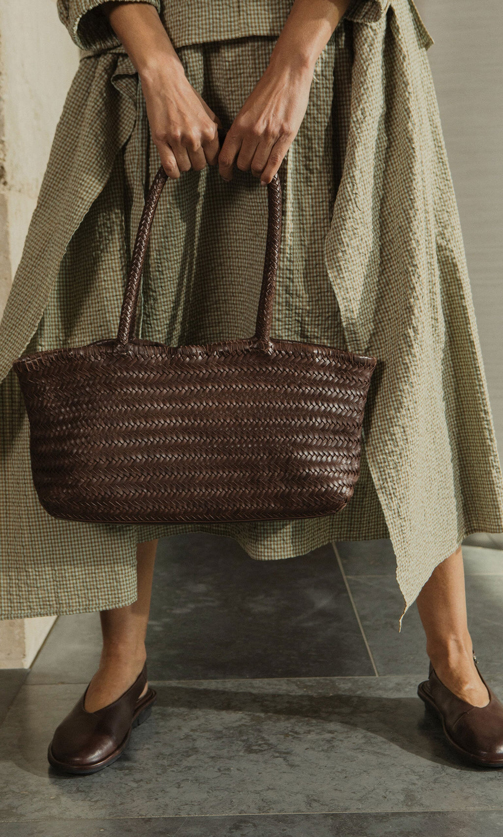Plümo- Person in a textured green dress holding the Inga Plait Bag, wearing brown flats on a tiled floor.