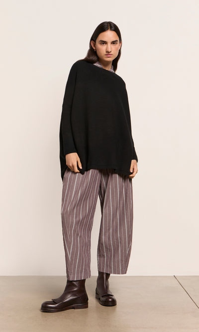 Plümo  Person in black sweater, Butoni Pants - Licorice, and brown boots stands against a beige background.