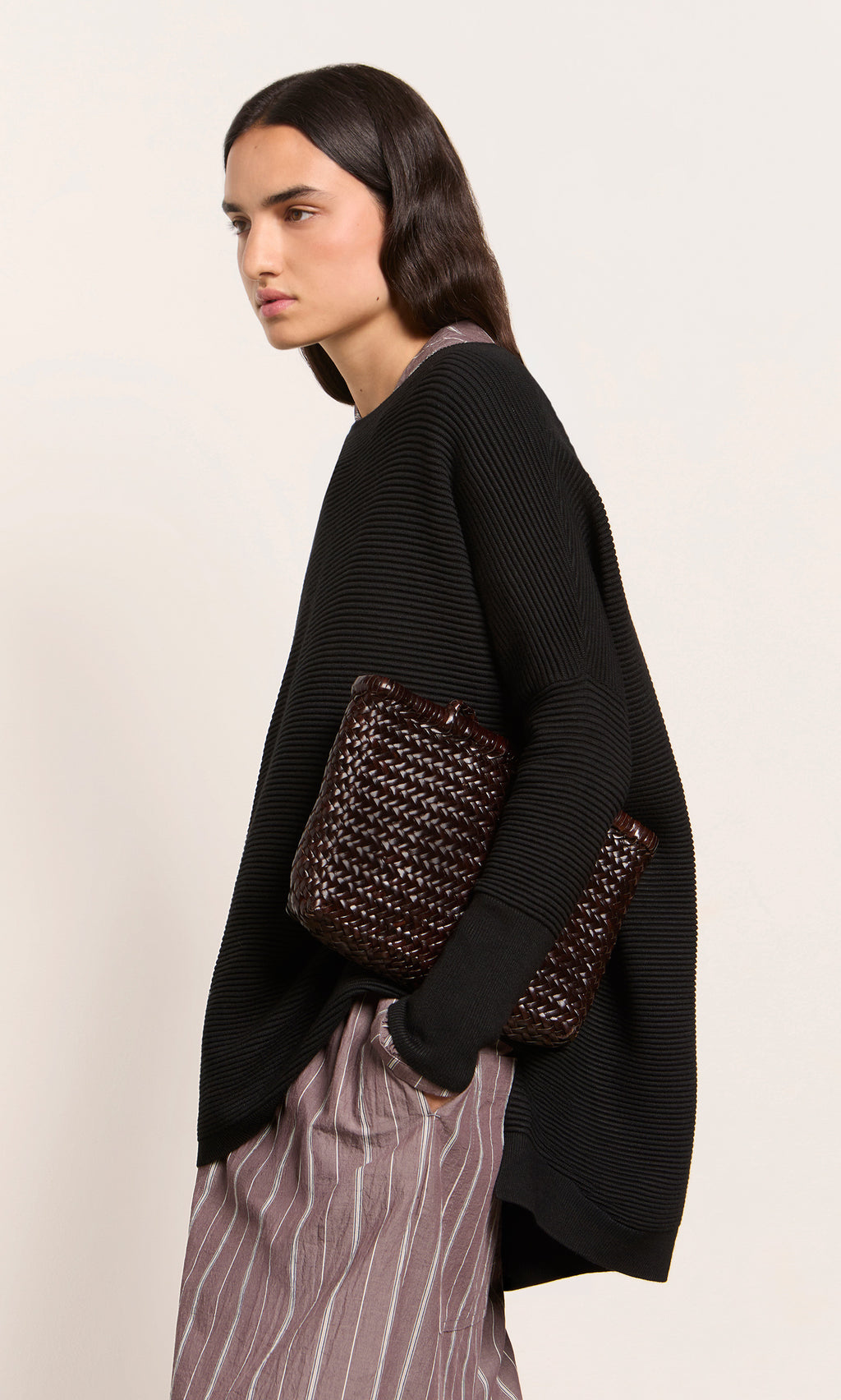 Plümo  Woman wears the Jack Sweater - Black with striped pants, holding a woven clutch against a plain background.