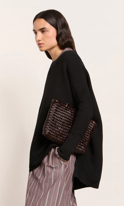 Plümo  Woman wears the Jack Sweater - Black with striped pants, holding a woven clutch against a plain background.