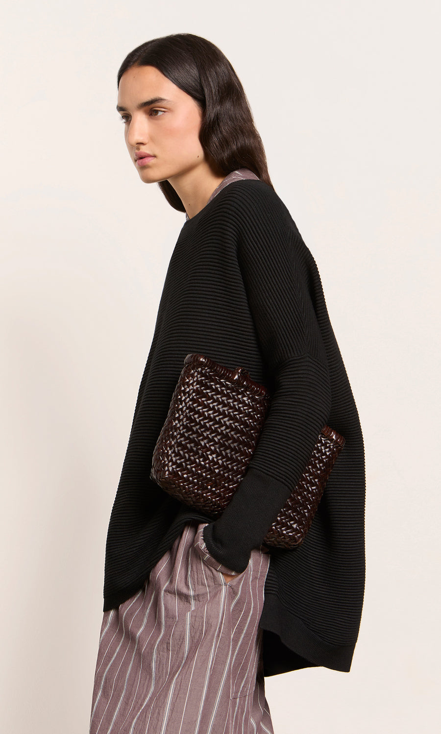 Plümo  Woman wears the Jack Sweater - Black with striped pants, holding a woven clutch against a plain background.
