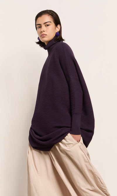 Plümo  Person wears Longer Jack Rollneck - Lupin and beige skirt, standing against a plain light background.