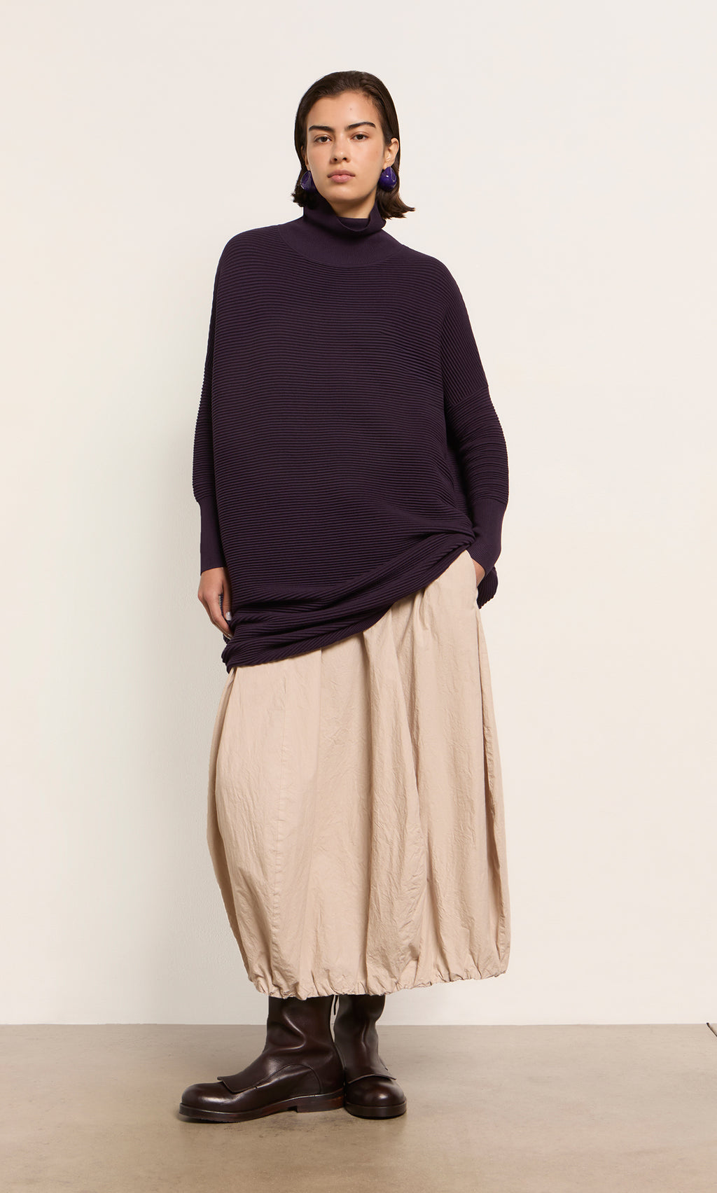 Plümo  Person in the Longer Jack Rollneck - Lupin, beige balloon skirt, and brown boots stands by a light wall.
