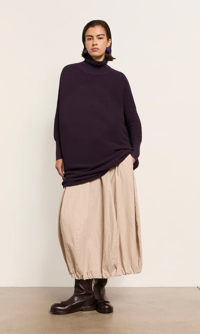 Plümo  Person in the Longer Jack Rollneck - Lupin, beige balloon skirt, and brown boots stands by a light wall.