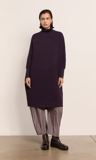 Plümo  Woman in a Longer Jack Rollneck - Lupin dress over gray striped pants, dark boots, stands by a light wall.