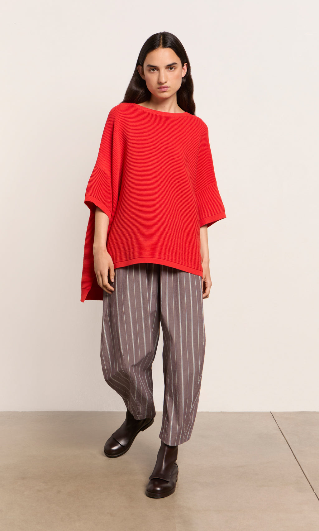 Plümo  Person wearing a lava red sweater, gray pinstriped pants, and black boots stands against a plain light background.
