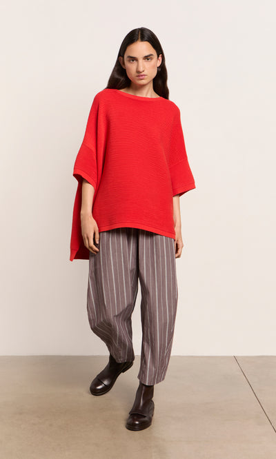 Plümo  Person wearing a lava red sweater, gray pinstriped pants, and black boots stands against a plain light background.