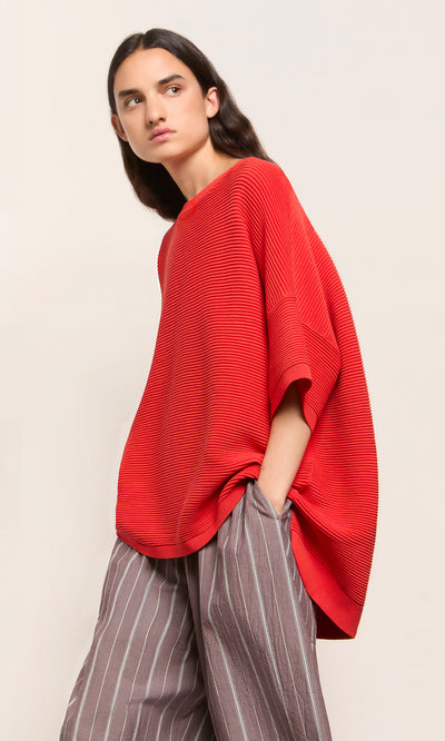 Plümo  Person in the Jack Sweater Short Sleeve - Lava stands sideways with a hand in pocket, wearing gray striped pants.