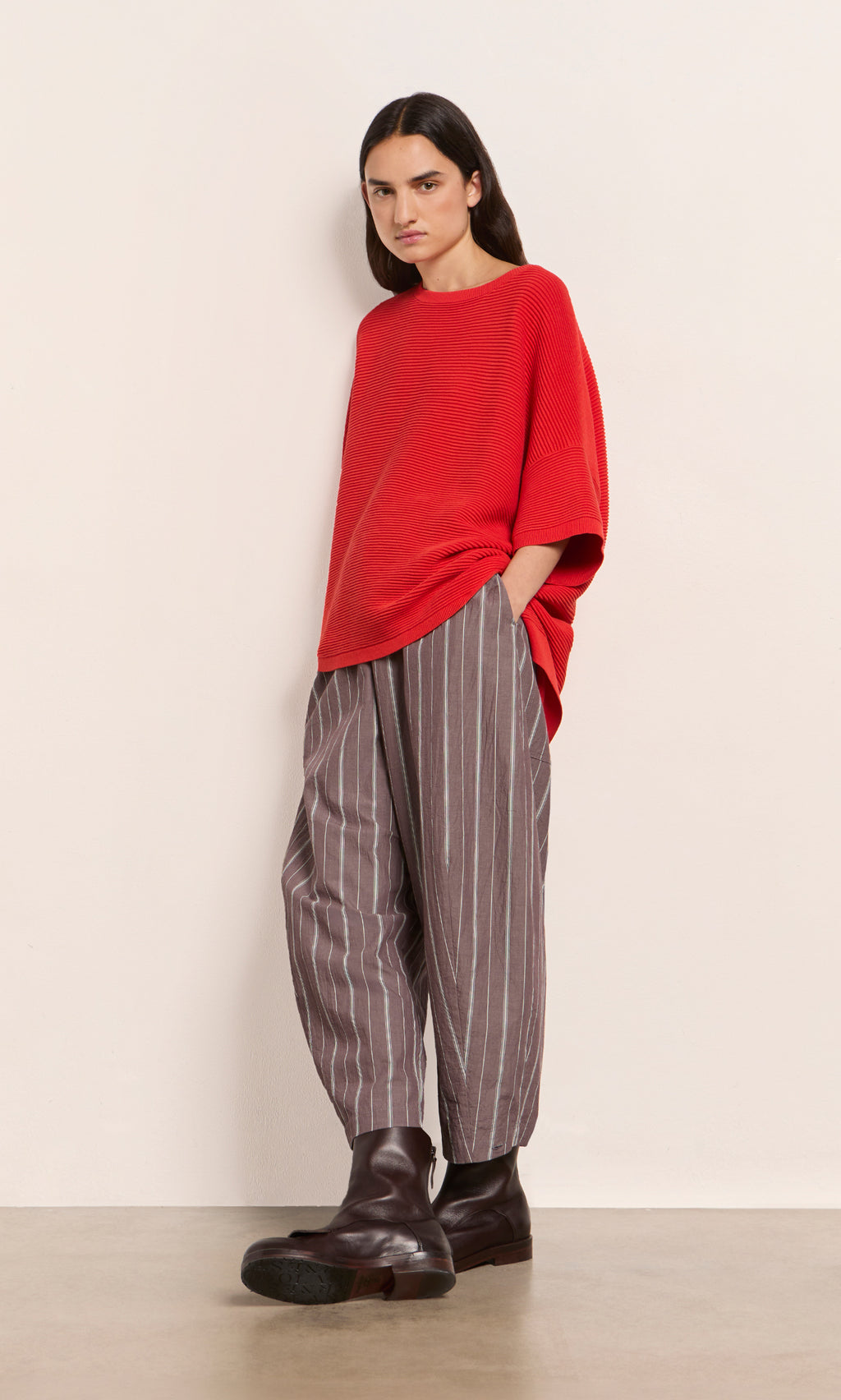 Plümo  Person wears a loose Jack Sweater Short Sleeve - Lava, striped gray pants, and dark boots by a plain light wall.