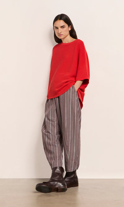 Plümo  Person wears a loose Jack Sweater Short Sleeve - Lava, striped gray pants, and dark boots by a plain light wall.