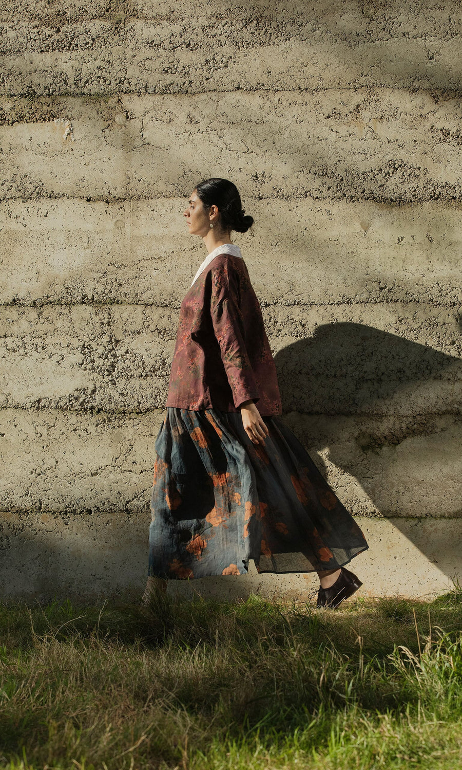 Plümo- A woman in the Jia Wrap Top and a patterned skirt walks outdoors by a textured concrete wall in sunlight.