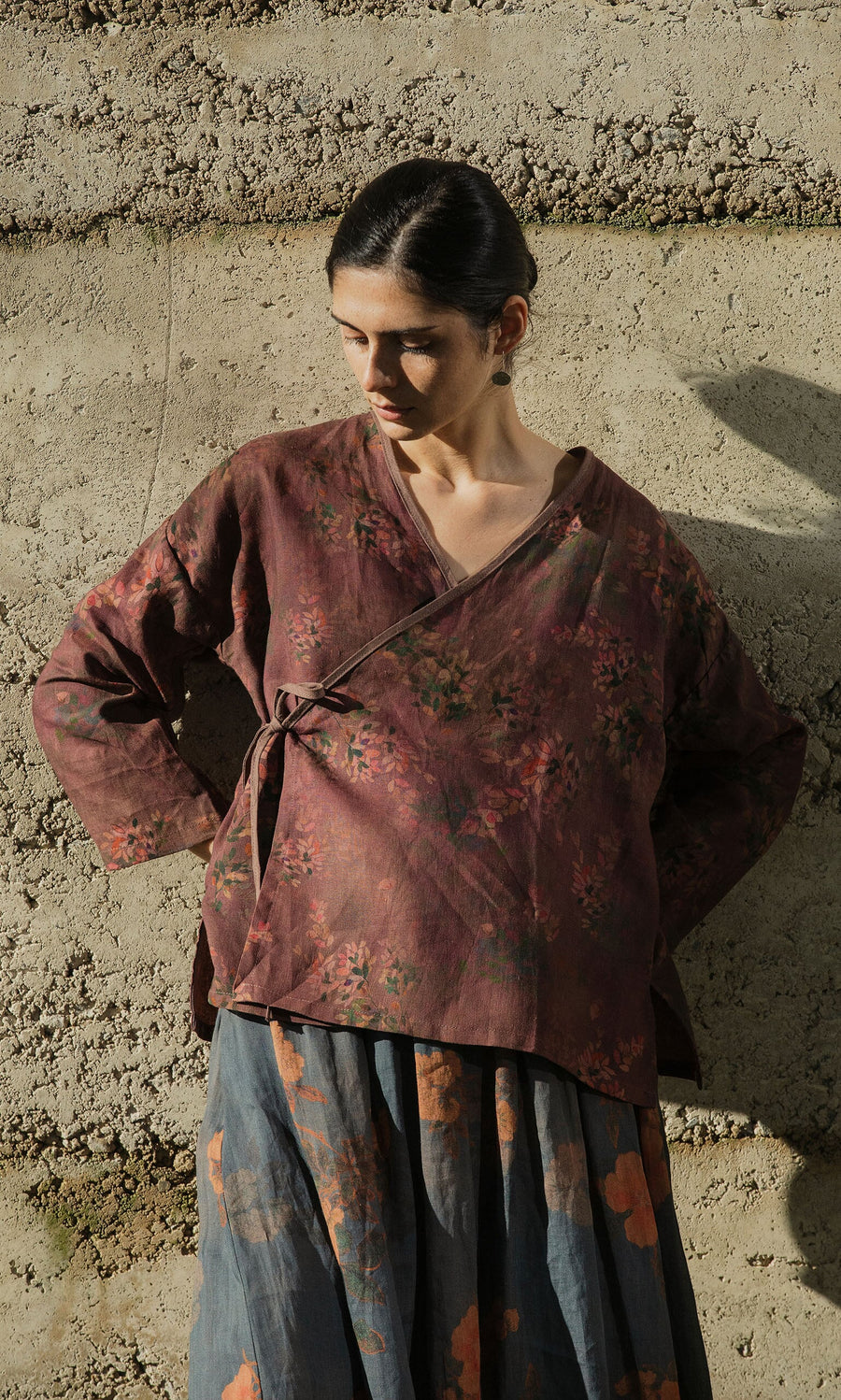 Plümo- Woman wears the Jia Wrap Top, standing against a concrete wall with hands on hips, looking down.
