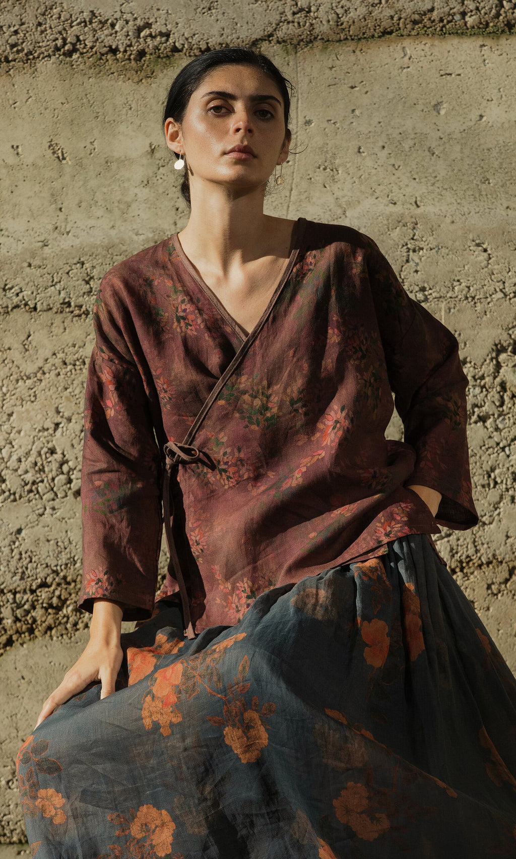 Plümo- A woman poses outdoors in the Jia Wrap Top and skirt, sunlight casting shadows against a textured concrete wall.