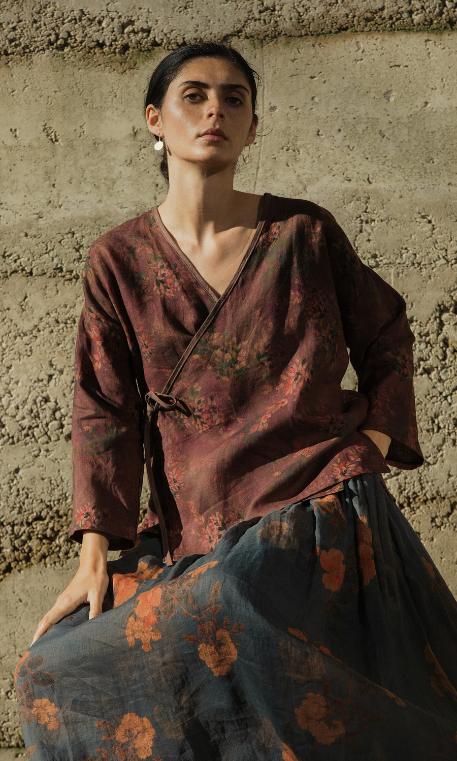 Plümo- A woman poses outdoors in the Jia Wrap Top and skirt, sunlight casting shadows against a textured concrete wall.