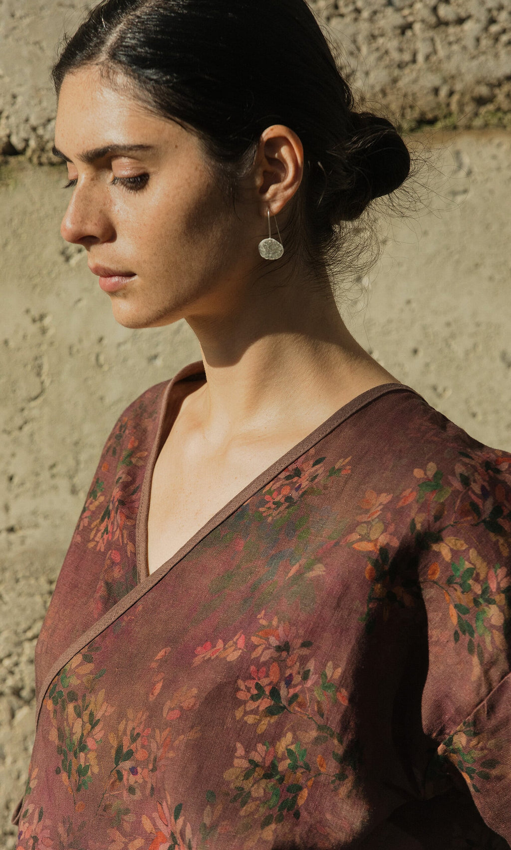 Plümo- Woman with dark hair in a bun wears the Jia Wrap Top and round earring, standing in sunlight.