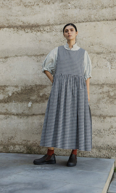 Plümo- A person in the Apron Dress - Gingham with spacious pockets stands against a textured wall, looking upward.
