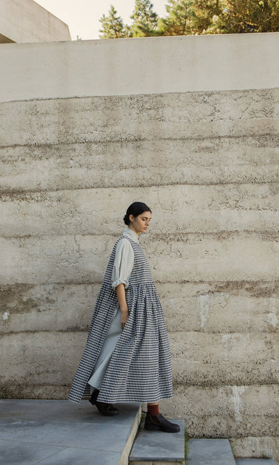 Plümo- Someone in the Apron Dress - Gingham and boots walks down concrete steps next to a textured beige outdoor wall.