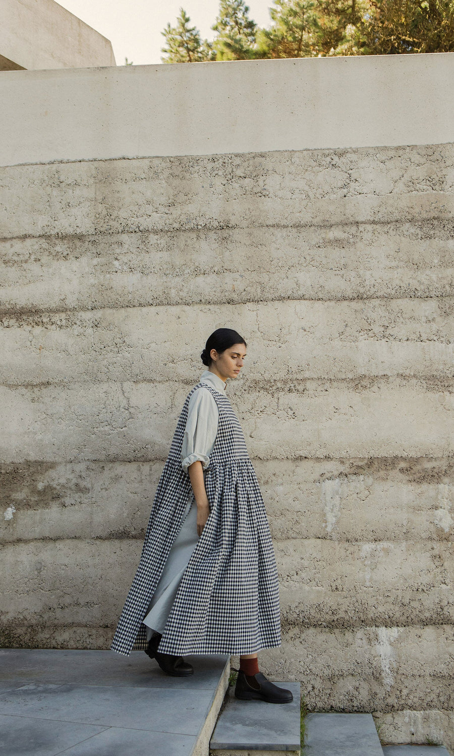 Plümo- Someone in the Apron Dress - Gingham and boots walks down concrete steps next to a textured beige outdoor wall.