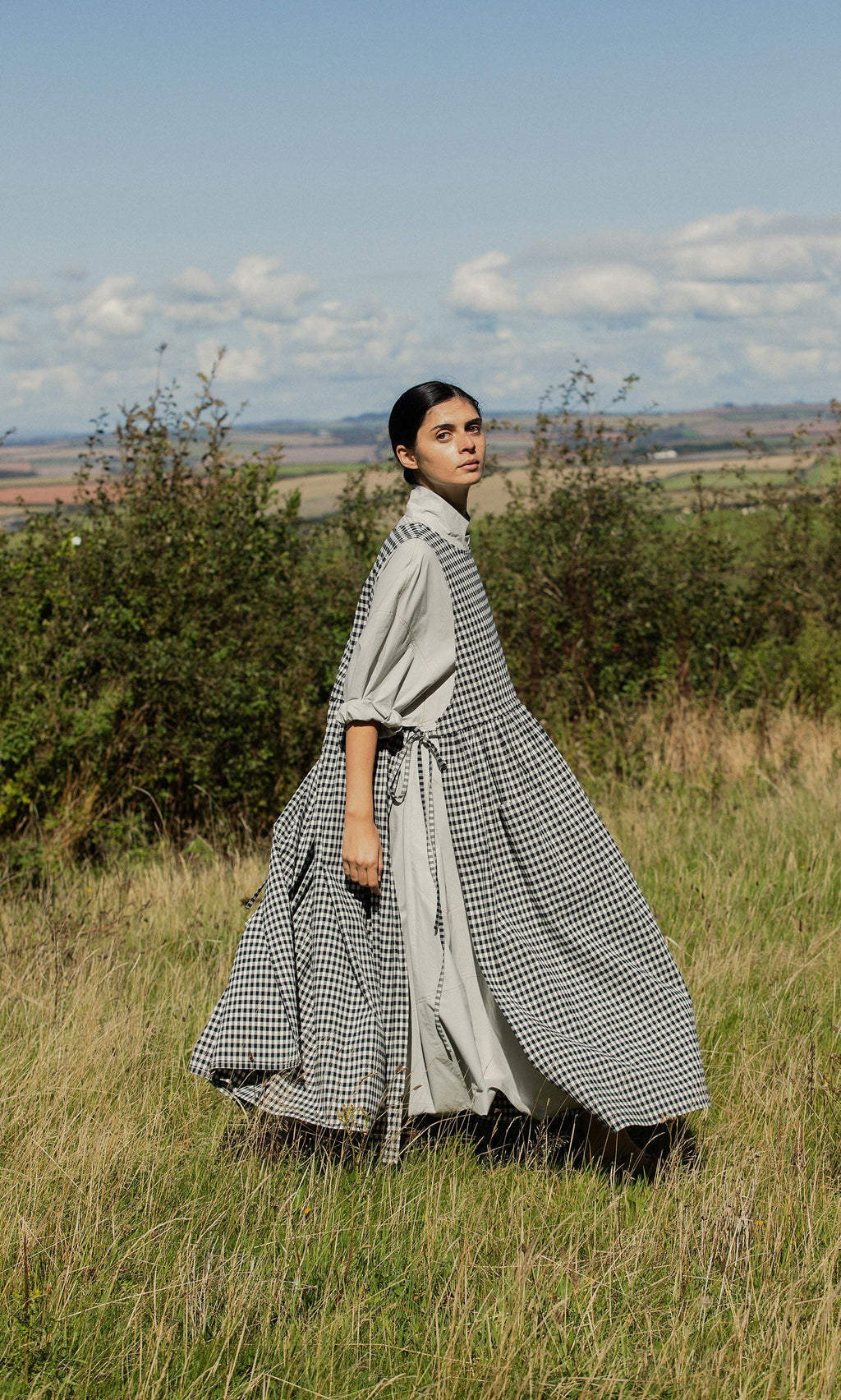 Plümo- A woman in the Apron Dress - Gingham walks through tall grass with hills and a partly cloudy sky behind her.