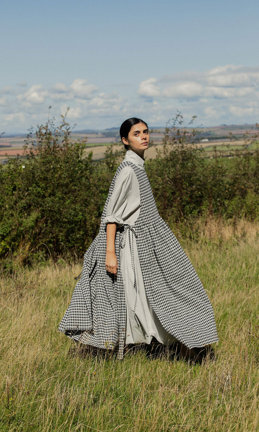 Plümo- A woman in the Apron Dress - Gingham walks through tall grass with hills and a partly cloudy sky behind her.